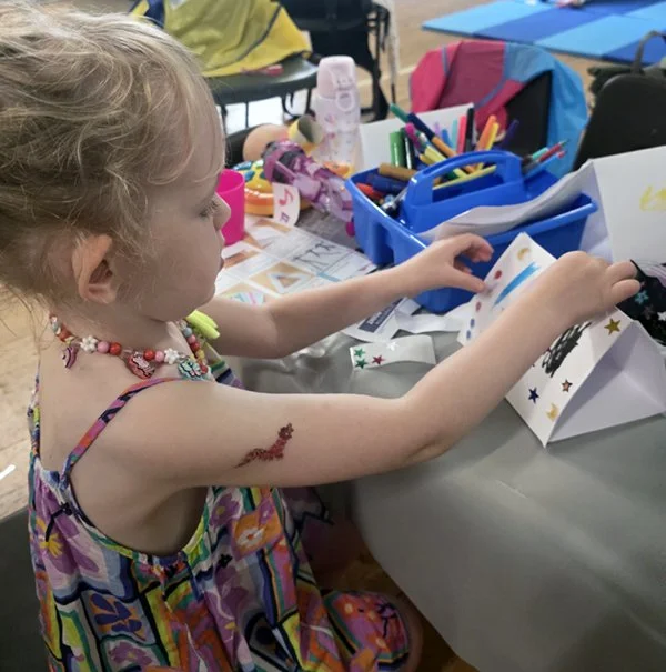 A young girl with a temporary arm tattoo and a colorful dress is sitting at a table, engaging with craft supplies such as paper, markers, and stickers in a room decorated for a celebration or arts and crafts activity.