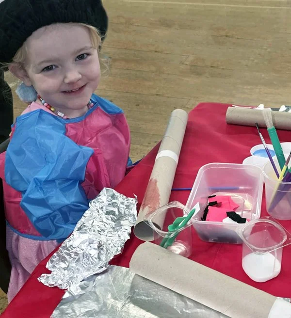 A young girl sitting at a table covered with craft supplies, including cardboard tubes, aluminum foil, and colored paper, smiling at the camera.
