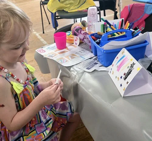 A young girl with blonde hair sitting at a table with art supplies, including markers, colored paper, and drawings, in a room with a wooden floor.