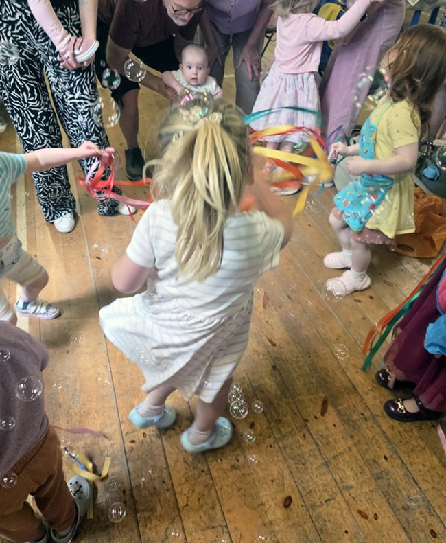 Children and adults playing with bubbles and ribbons at a party indoors, with a wooden floor.