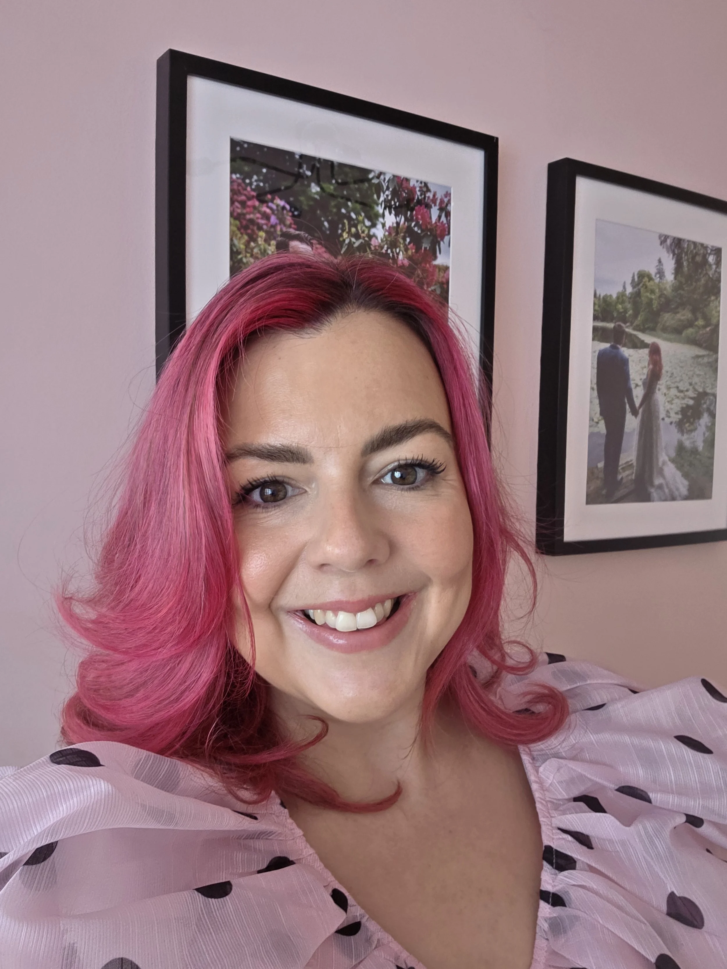 A woman with bright pink hair smiling in front of framed wedding photos on a pink wall.
