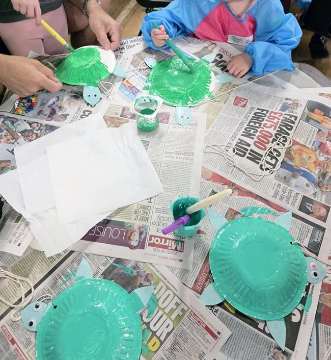 Children painting turtle-shaped craft project with green paint, using brushes, on a table.
