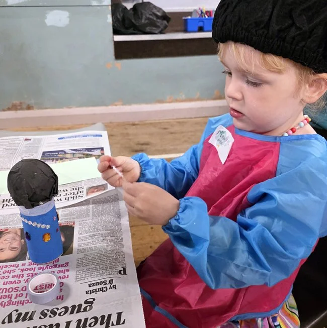 A young child with blonde hair wearing a black hat and a red and blue smock, sitting at a table. The child is holding a small object and appears to be engaged in a craft activity. Making a microphone for festival themed crafts.