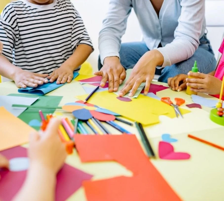 Children and an adult engaging in arts and crafts with colorful paper, scissors, glue, and markers.