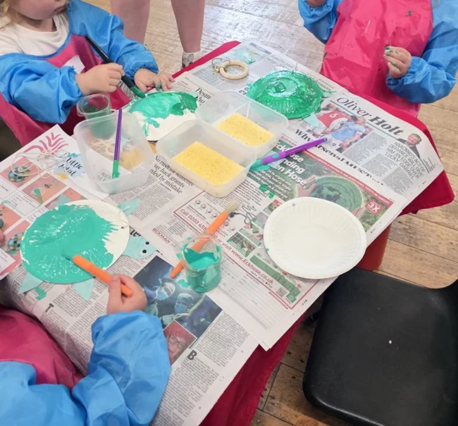 Children painting with green paint at a table, jars of yellow textured substance, and a paper plate.