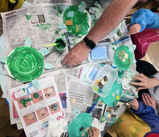 Children and adult making turtle-shaped crafts with painted paper plates, paper parts, and glue on a table.