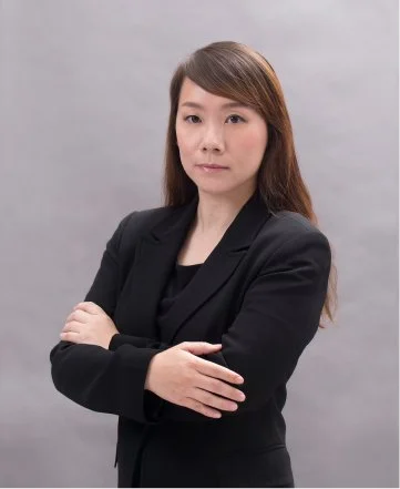 A woman with long brown hair wearing a black blazer and black top, posing with her arms crossed in front of a gray background.