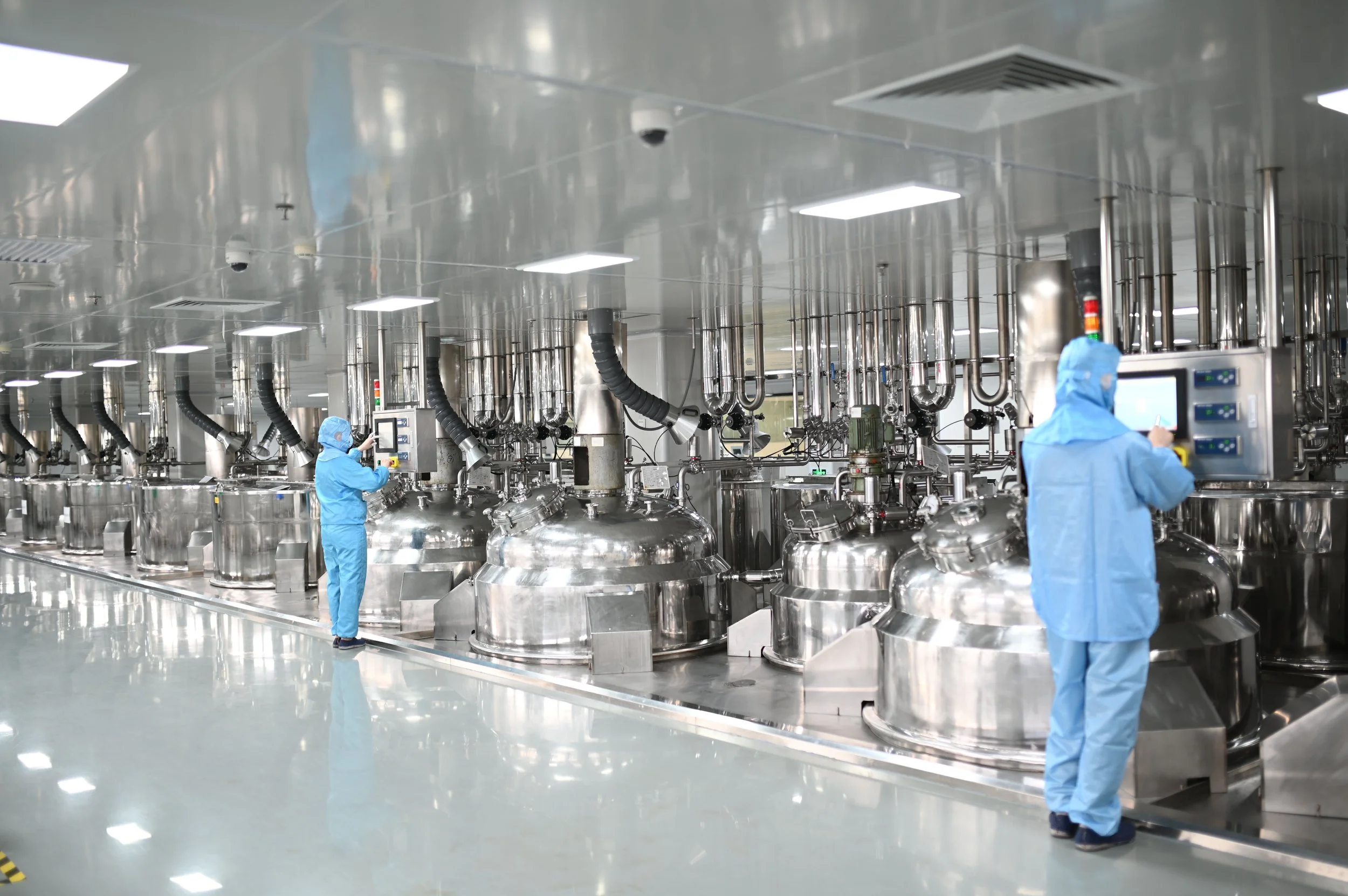 Workers in blue protective suits operating machinery in a clean, stainless steel pharmaceutical or biotech manufacturing facility.