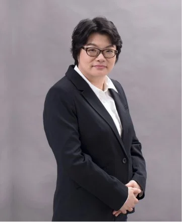 A woman in a black blazer and glasses posing against a plain gray background.