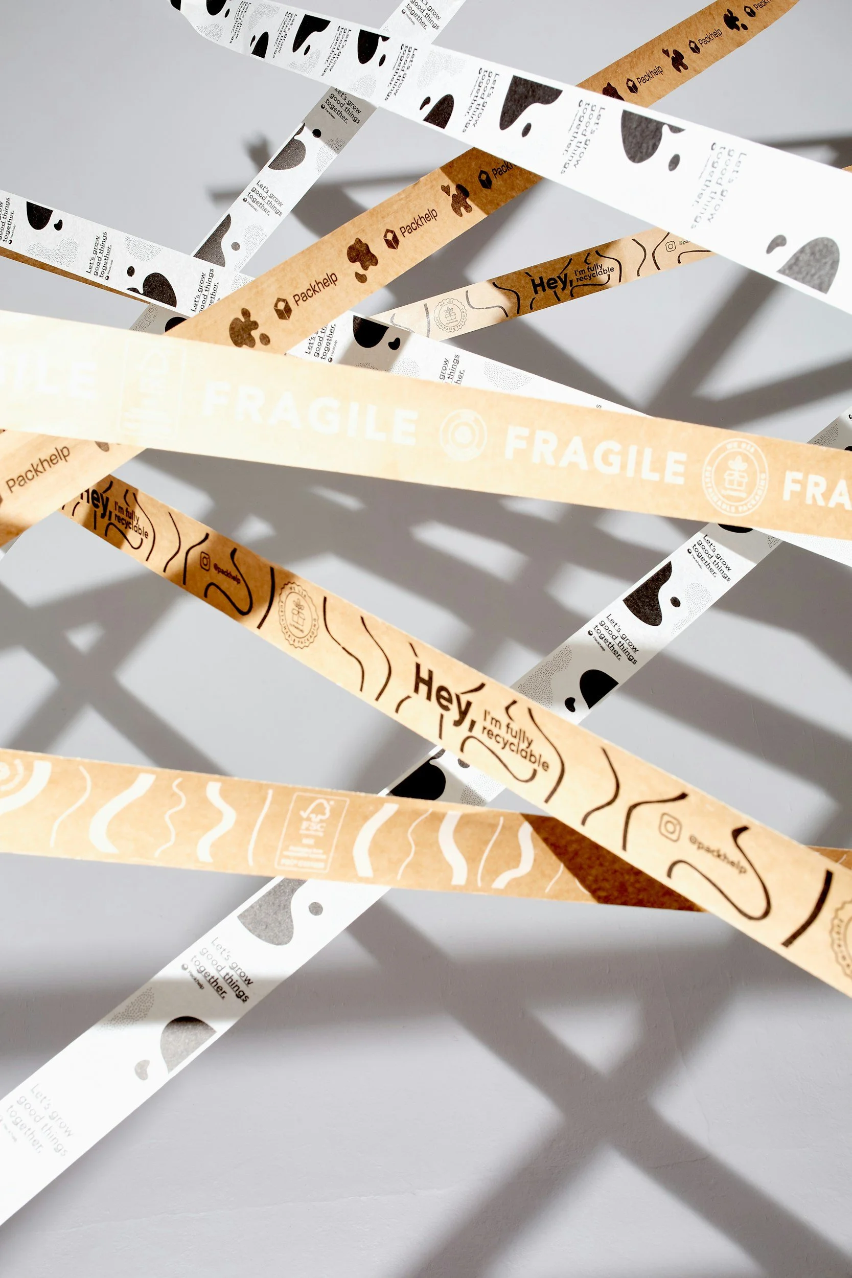 Various recyclable paper strips with printed symbols and messages, including 'Fragile,' 'Hey, I'm fully recyclable,' and icons indicating recycling, placed on a white surface with shadows cast by the strips.