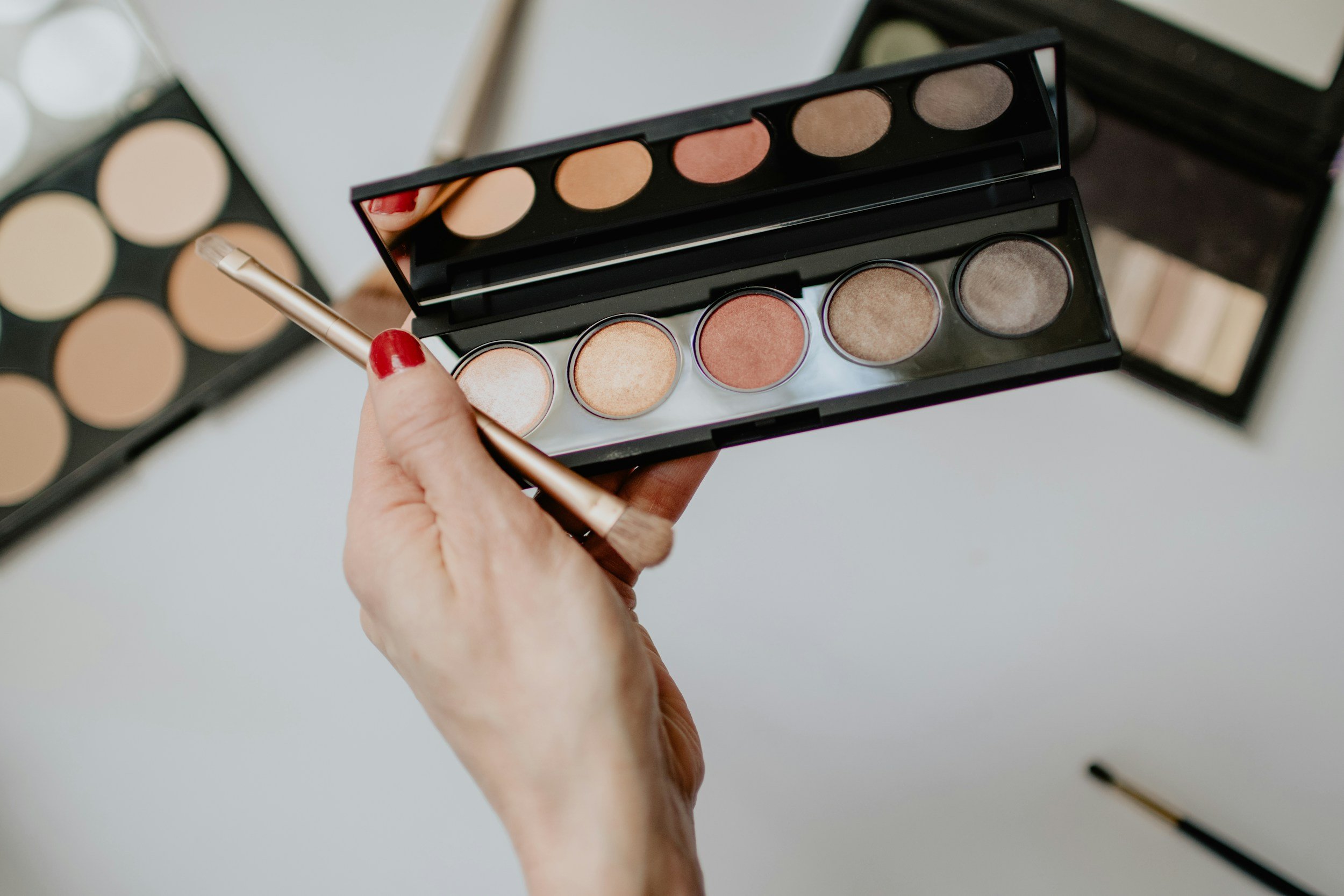 A person with red nail polish holding an eyeshadow palette with neutral shades, with a makeup brush. In the background, there are two additional eyeshadow palettes with similar shades.