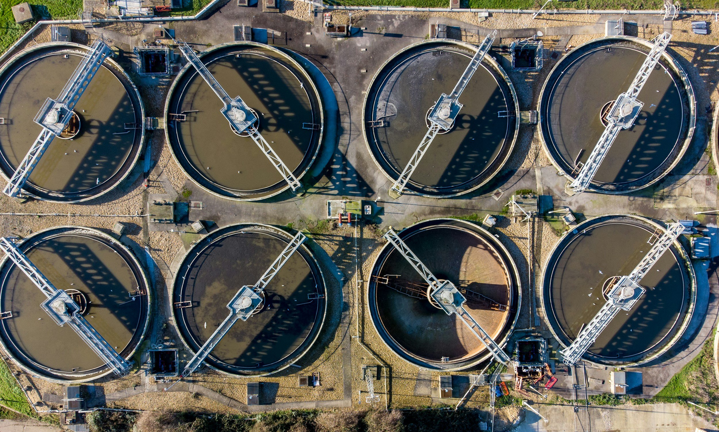 Aerial view of a wastewater treatment plant with circular clarifiers filled with water, connected by metal walkways and support structures.