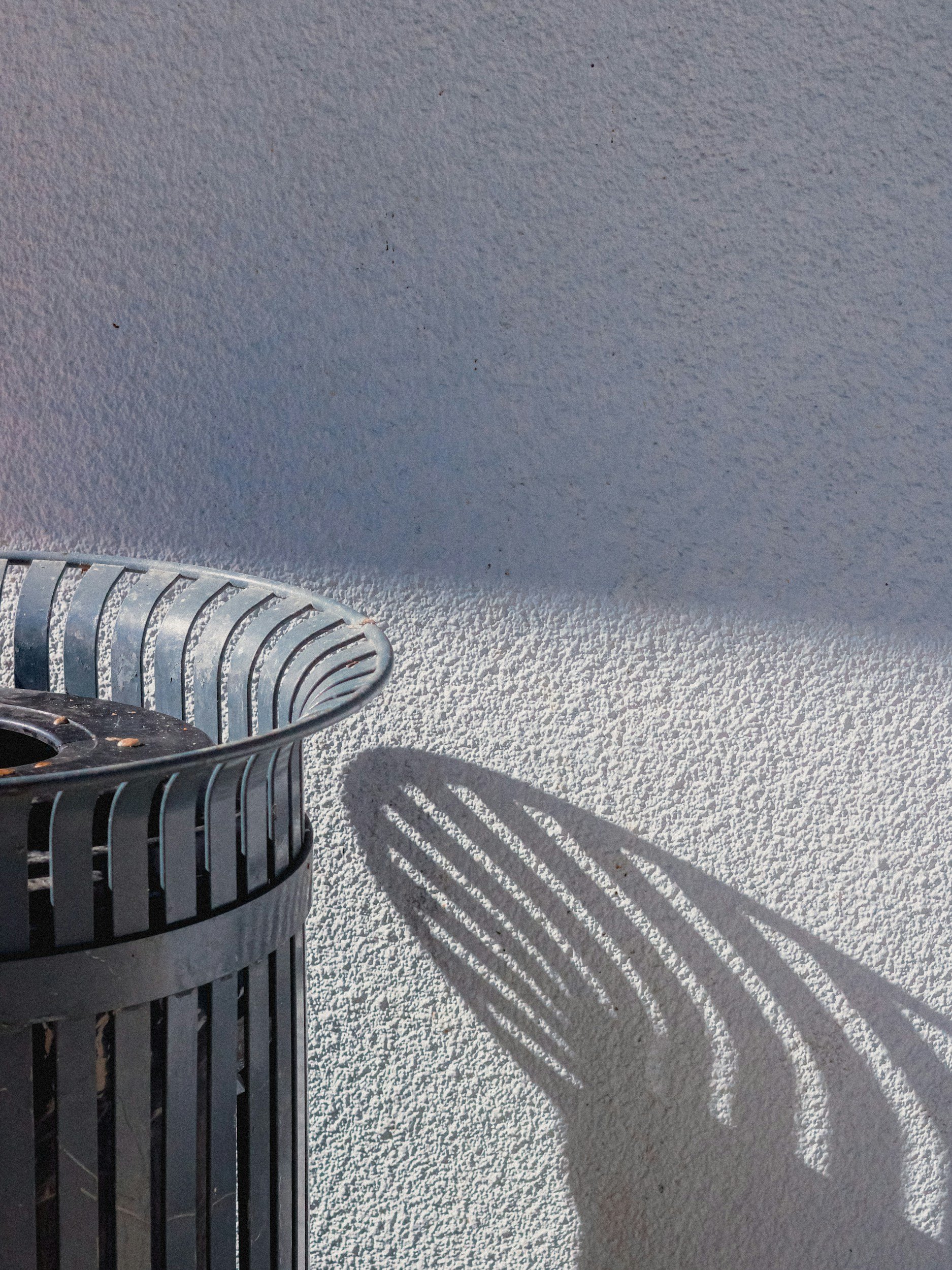 A metal trash can with vertical slats casting shadows on a textured white surface, possibly concrete, with a shadow of the trash can visible on the surface.