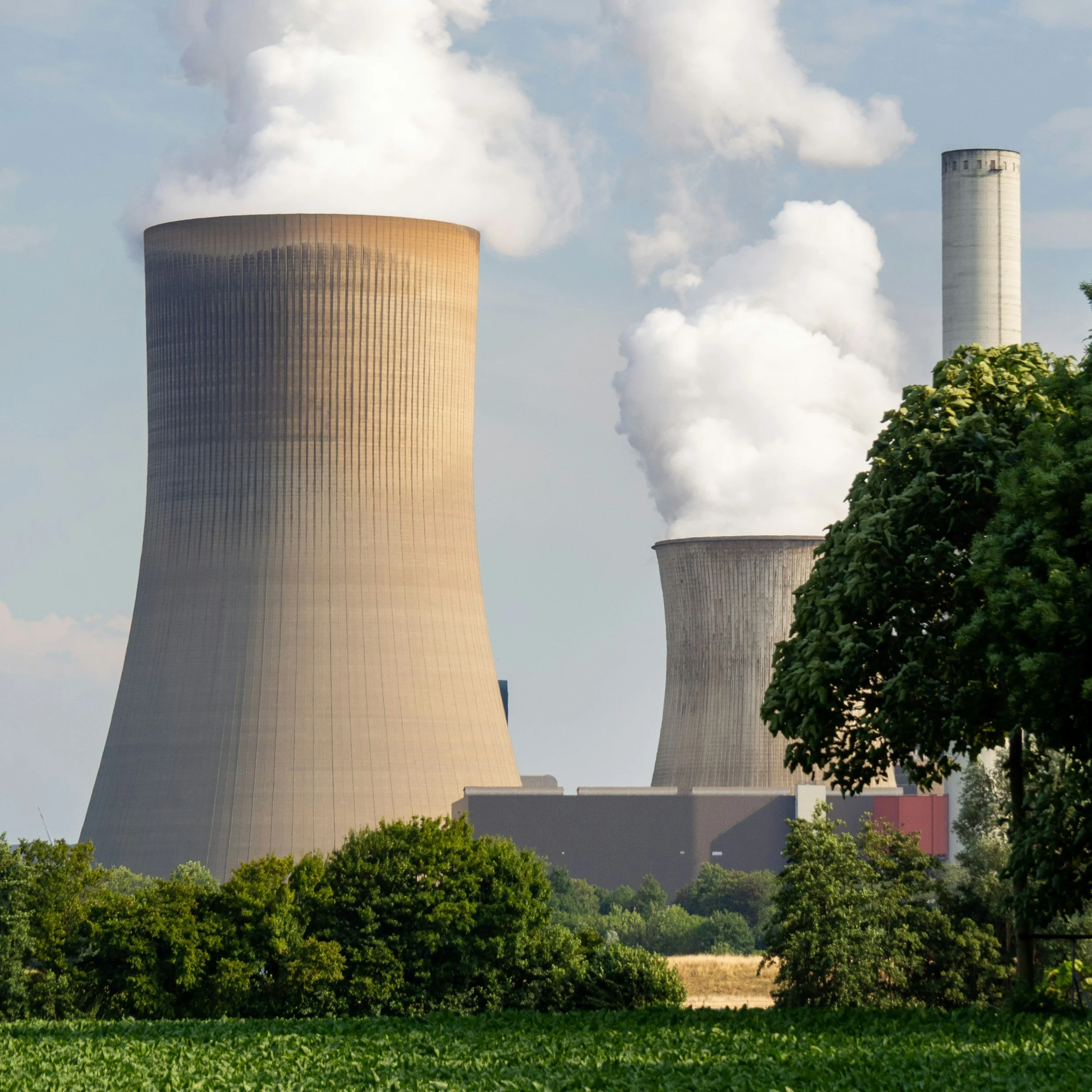 Nuclear power plant cooling towers emitting steam seen behind green trees and vegetation.