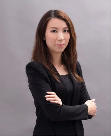 A woman with shoulder-length brown hair wearing a black blazer and black top, crossing her arms, standing against a neutral gray background.