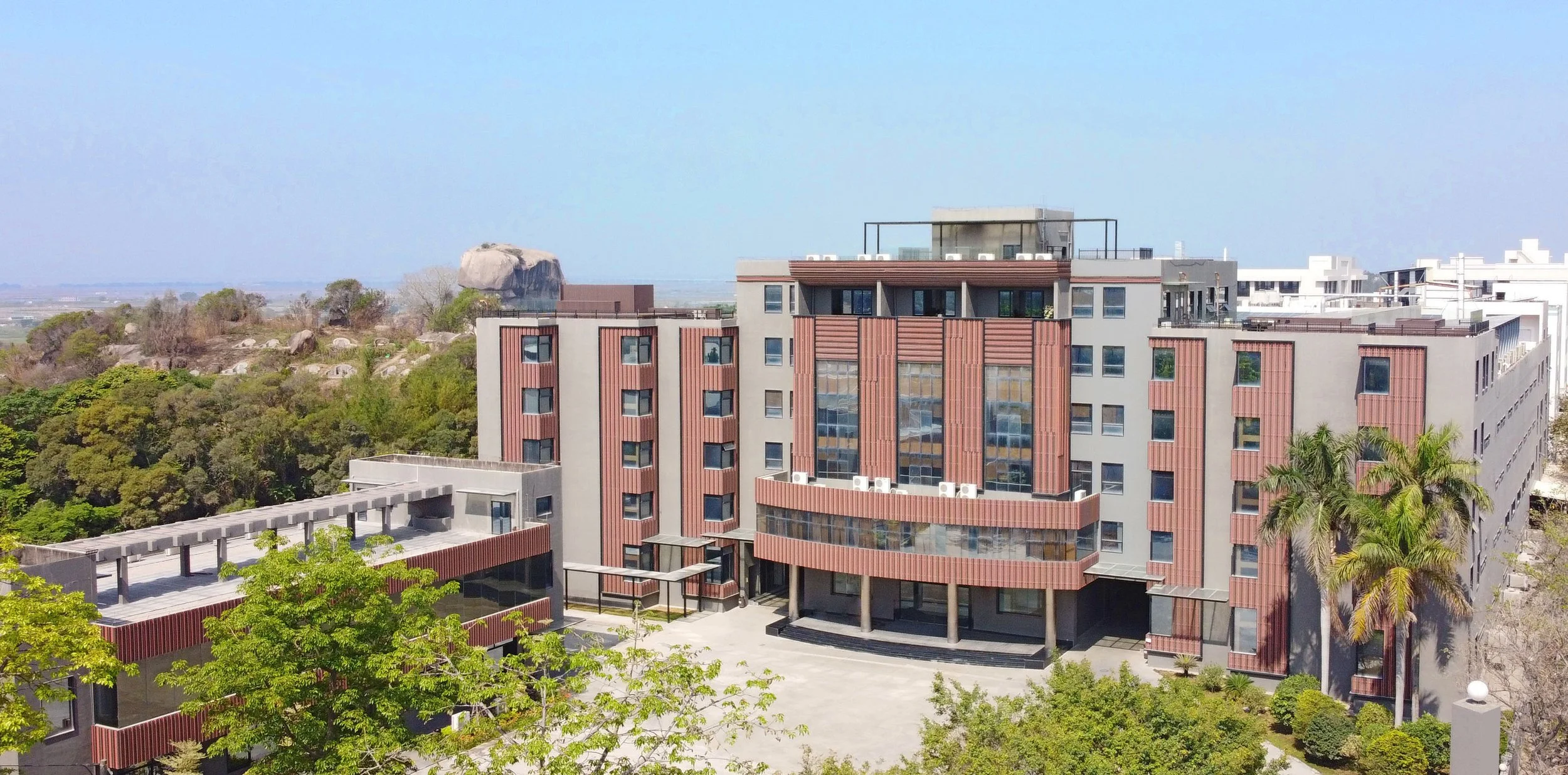 A modern multi-story building with reddish-brown vertical slats, surrounded by green trees and palm trees. The building features large windows, balconies, and a landscaped courtyard in front. In the background, there is lush greenery, rocky terrain, and a clear blue sky.
