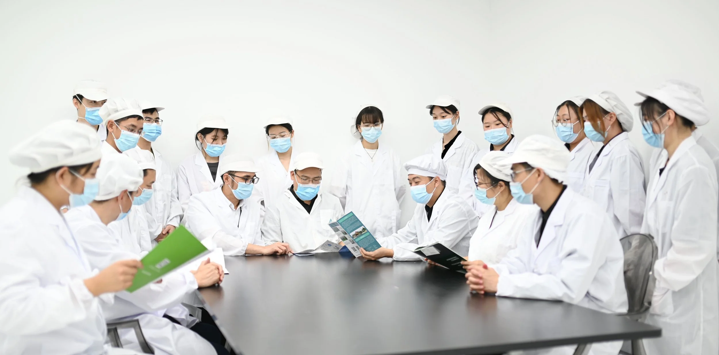 Group of people in white lab coats and masks gathered around a table, with some reading brochures, in a clean, white room.