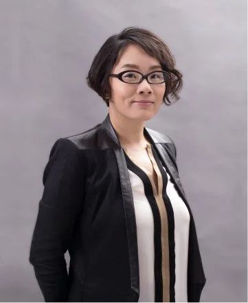 A woman with short dark hair, glasses, and a black blazer over a white blouse with black and tan vertical stripes, standing against a gray background.