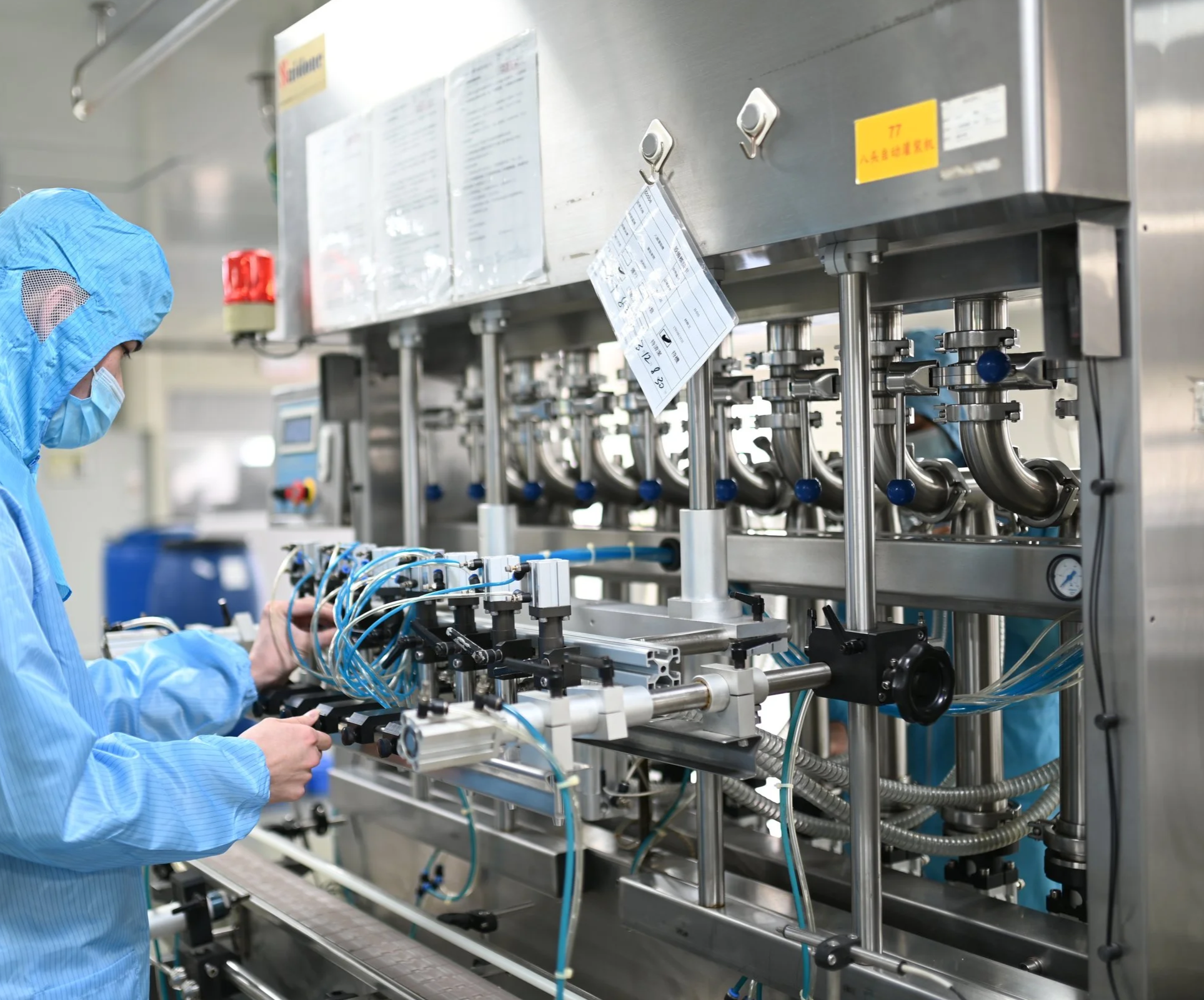 A worker in blue protective clothing, face mask, and hair cover operating industrial machinery with tubes, valves, and electronic controls in a clean manufacturing environment.