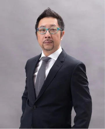 A man in a dark suit, white shirt, and tie standing against a gray background.