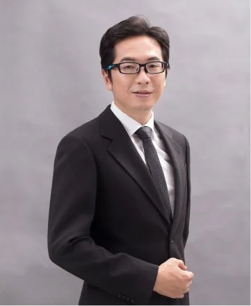 A man in a black suit, white shirt, and gray tie standing against a gray background.
