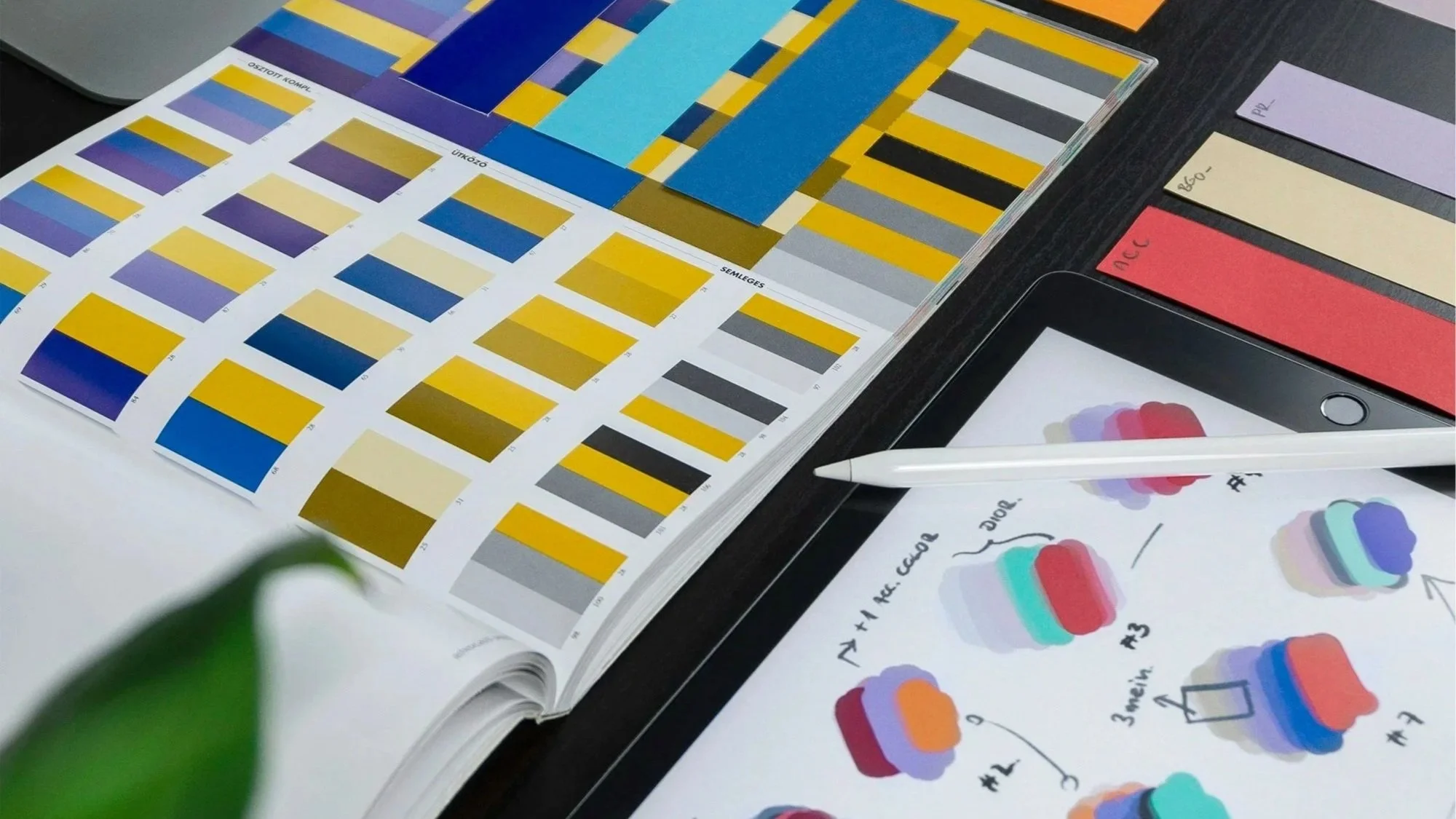 A color swatch book with various yellow, blue, purple, and gray shades, a printed sheet with color illustrations and labels, several color sample strips, a tablet, and a white pen on a black desk.