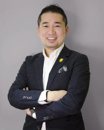 A smiling man in a dark suit with a white shirt, standing with arms crossed against a plain gray background.