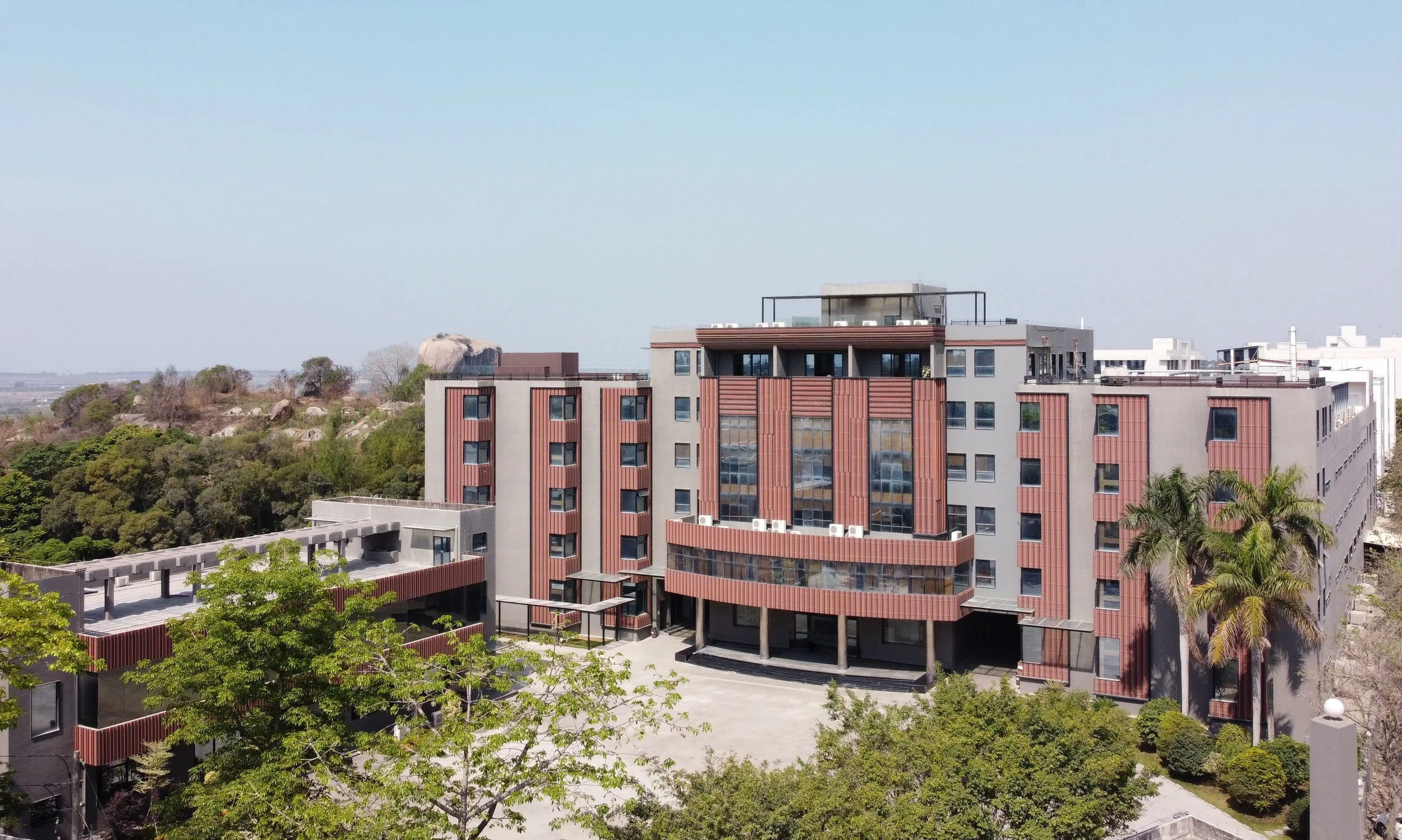 Modern multi-story residential or hotel building with red and gray exterior, surrounded by green trees and palm trees, under a clear blue sky.