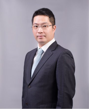 A man wearing glasses in a dark suit and tie, posing against a gray background.
