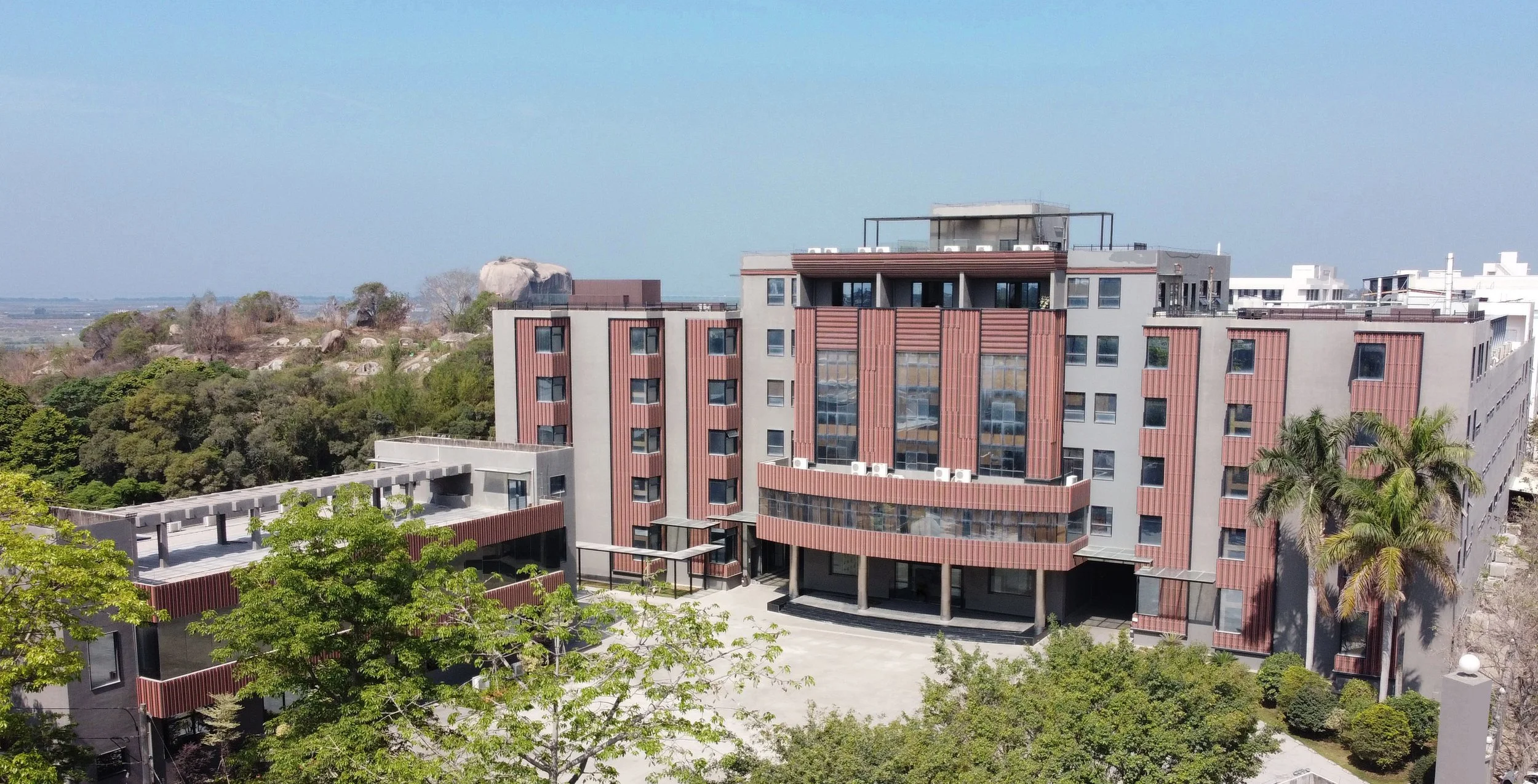 Modern multi-story residential or commercial building with large glass windows, surrounded by trees and greenery, under a clear blue sky.