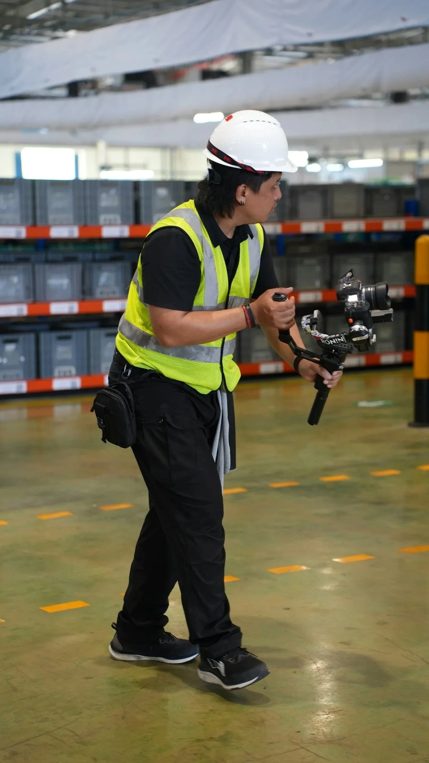 A person wearing a white safety helmet, a yellow reflective vest, and black pants is holding a camera stabilizer rig in a warehouse or industrial setting with shelves and boxes in the background.