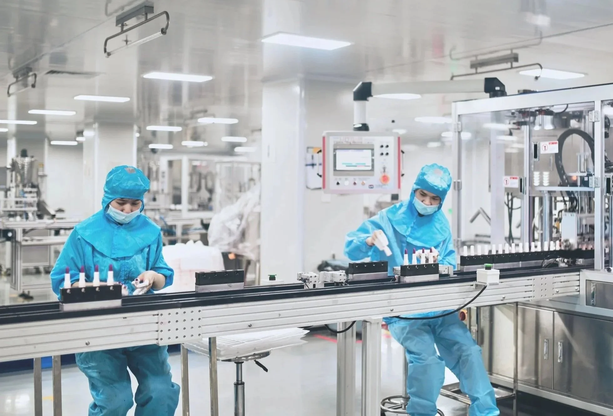 Two workers in blue protective suits and masks working on a production line in a clean manufacturing environment.