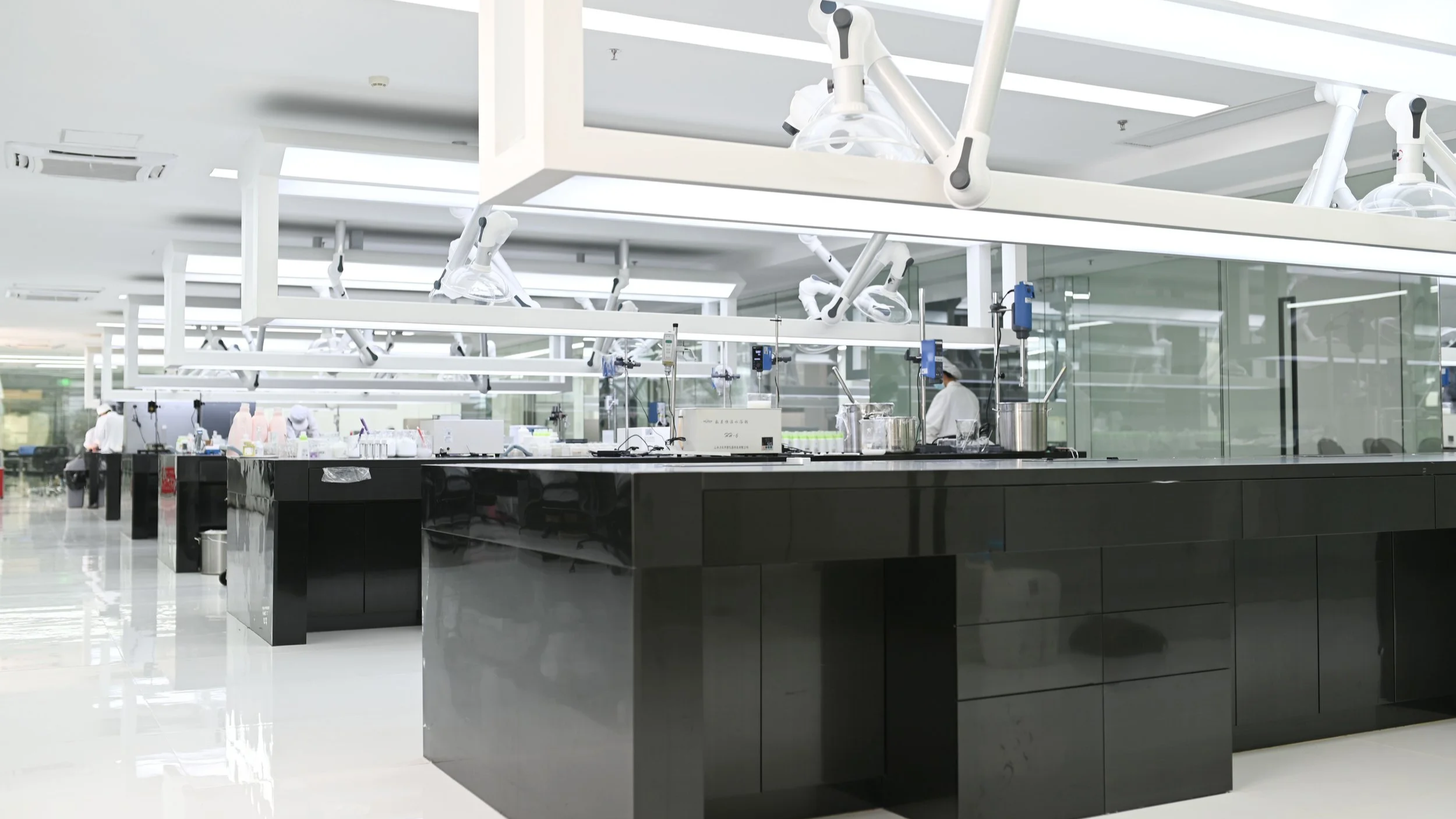 A modern medical laboratory with black countertops, robotic arms, and glass walls. Several employees in white lab coats are working at different stations in the background.