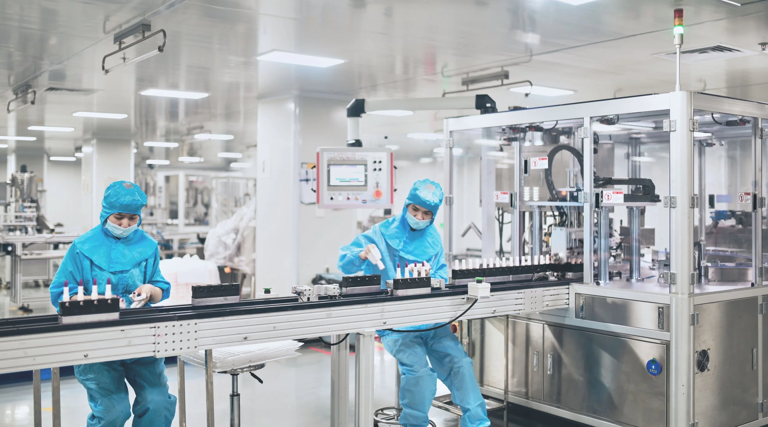Two workers in blue protective suits and masks operate machinery on a production line in an industrial manufacturing facility.