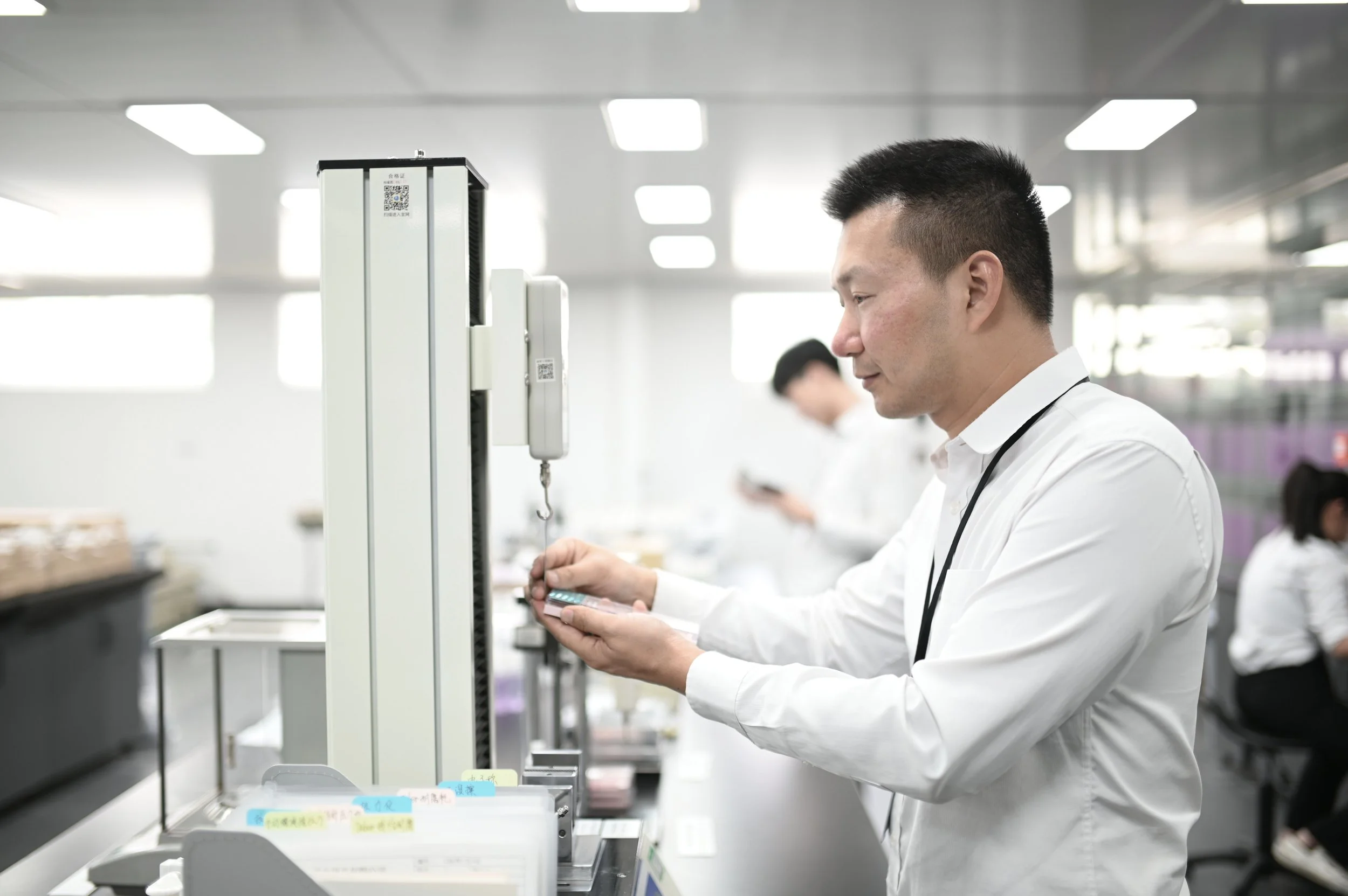 A man in a white shirt working in an industrial or laboratory setting, holding a phone, with other people in the background in a bright, modern workspace.