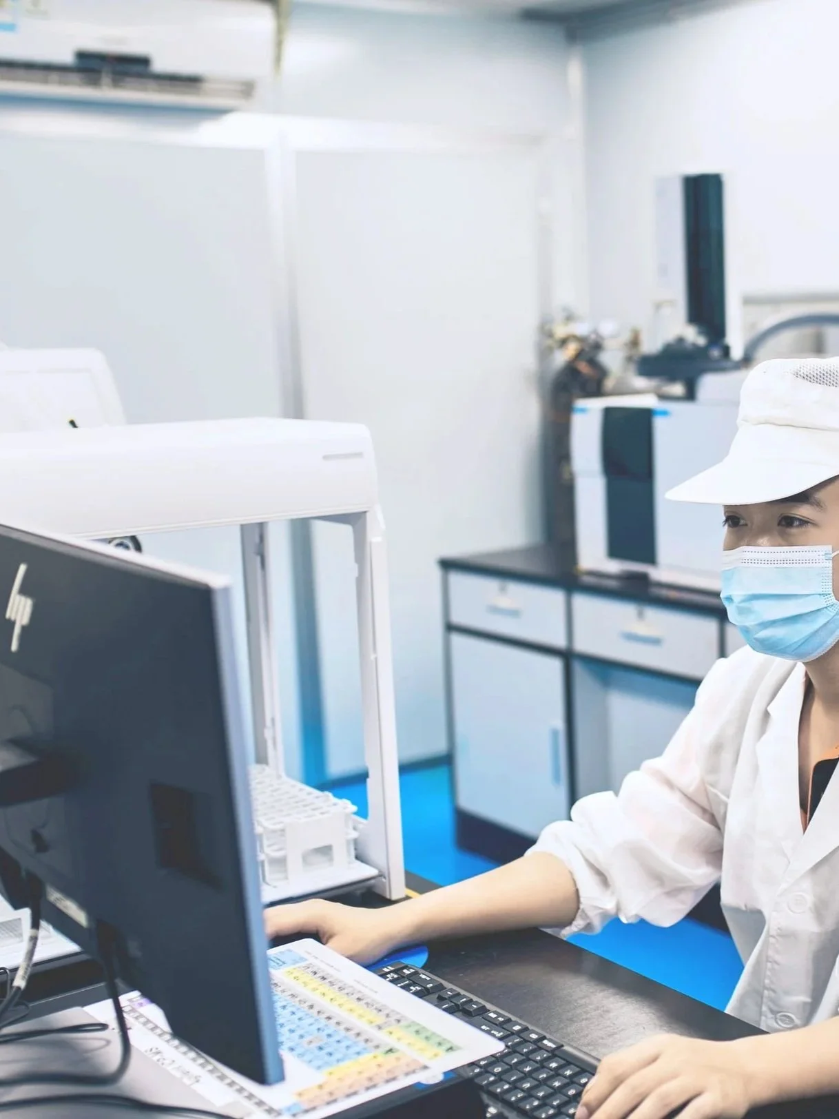 A healthcare worker in a white uniform, wearing a face mask and cap, working at a computer laboratory station with scientific equipment in a clinical or laboratory setting.