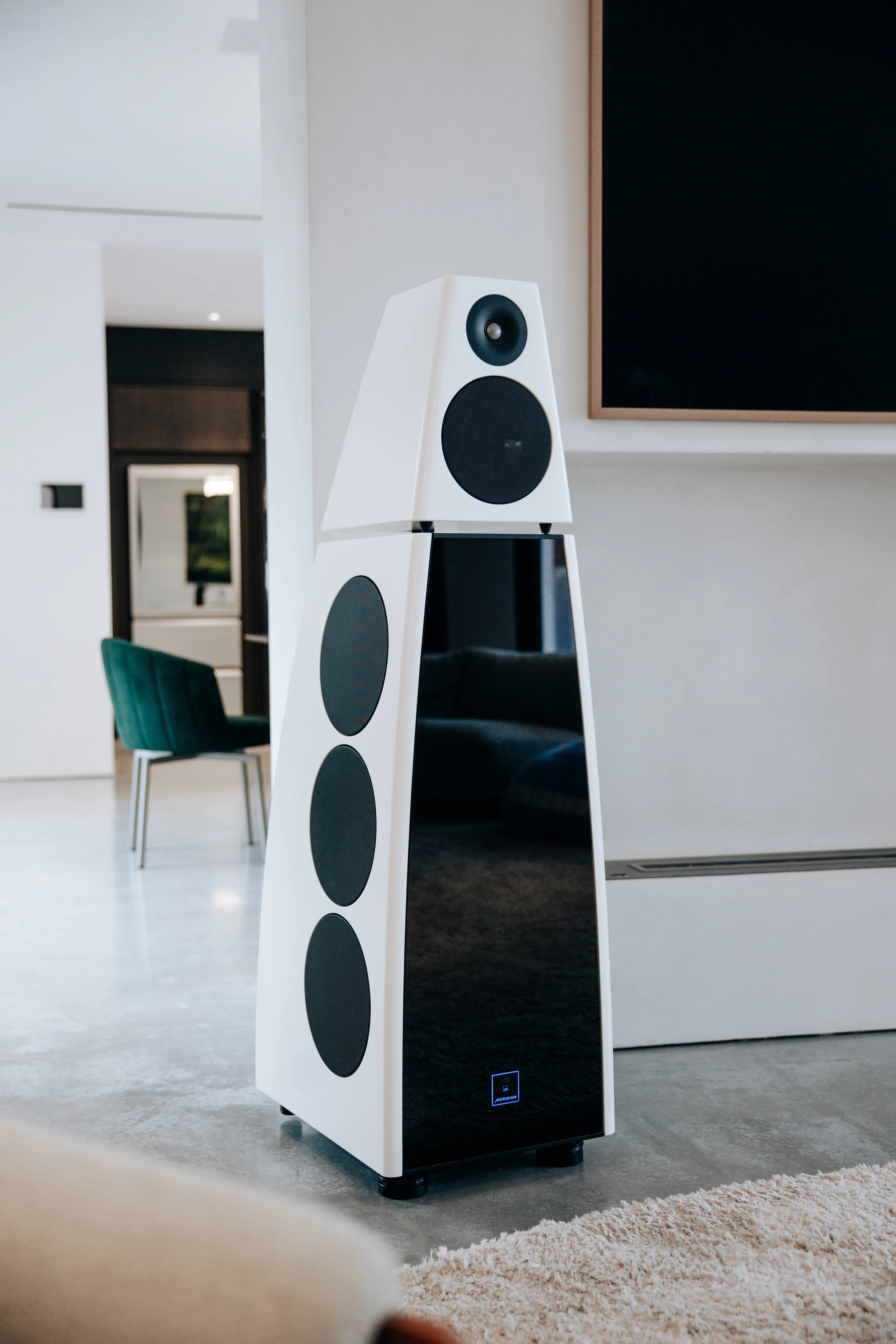 A large Meridian Audio floor-standing speaker with a white and black finish, positioned in a contemporary living room with a television and chair in the background.
