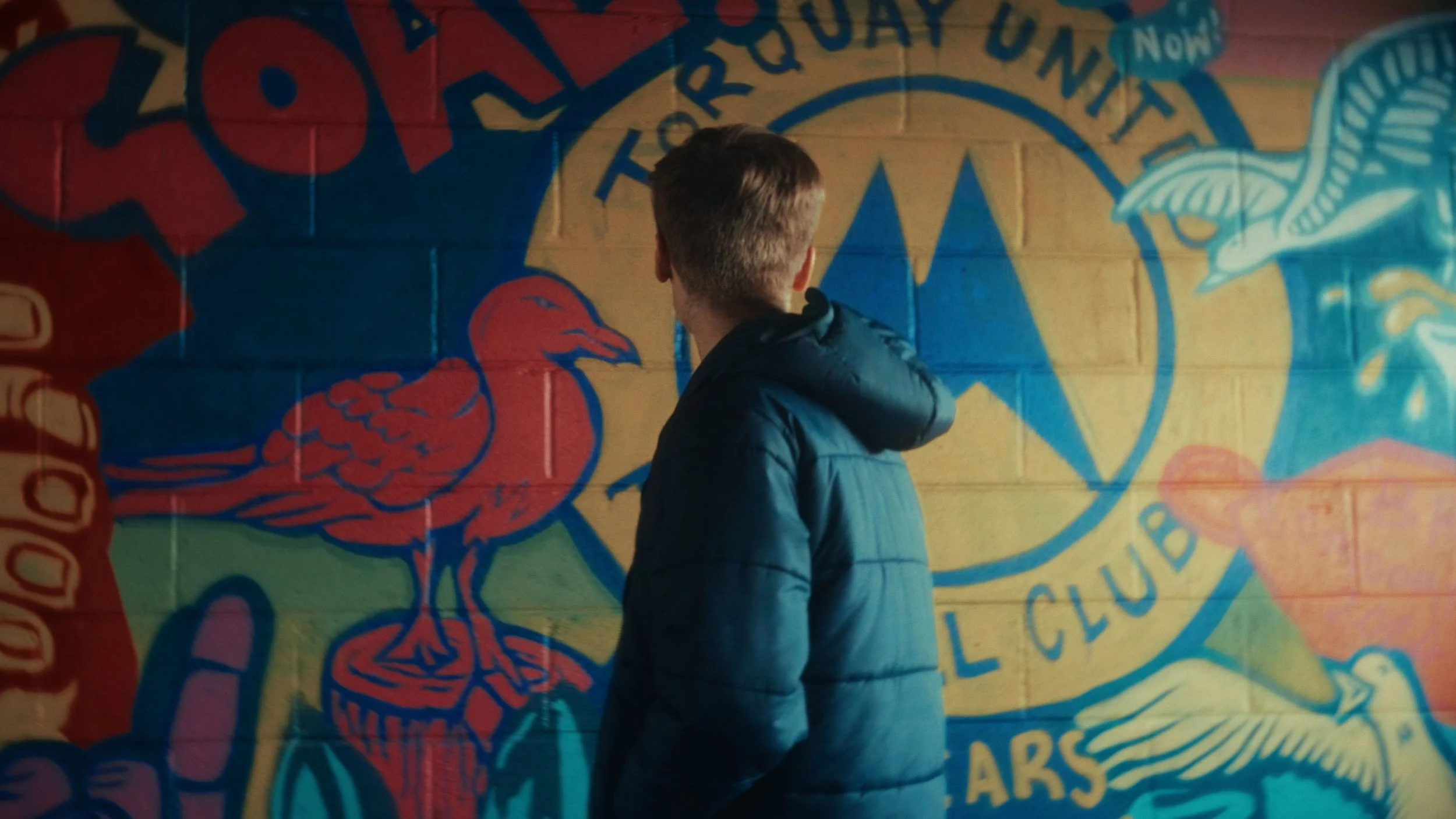 A person with short hair wearing a dark blue puffy jacket stands with their back to the camera, in front of a colourful mural with a Torquay United club logo