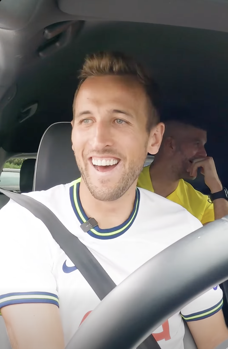 Harry Kane sitting in a car, one in the front seat smiling and looking outside, the other man in the back seat is talking