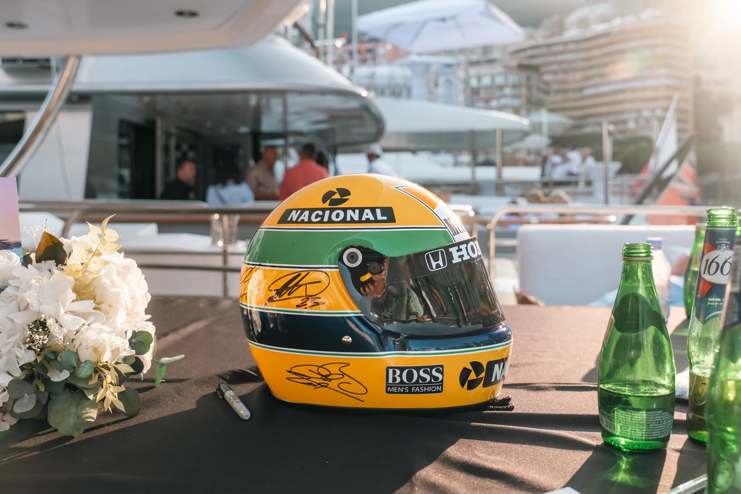 A replica Ayrton Senna racing helmet with yellow, green, and black colors, signed autographs, and sponsor logos including 'Nacional,' 'Honda,' and 'Boss' on a table with green bottles and flowers at a marina.