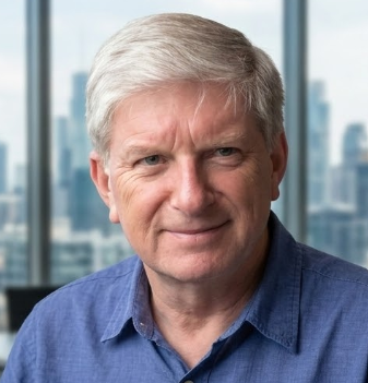 A security consultant and businessman with gray hair, wearing a blue shirt, smiling at the camera with city skyline in the background.