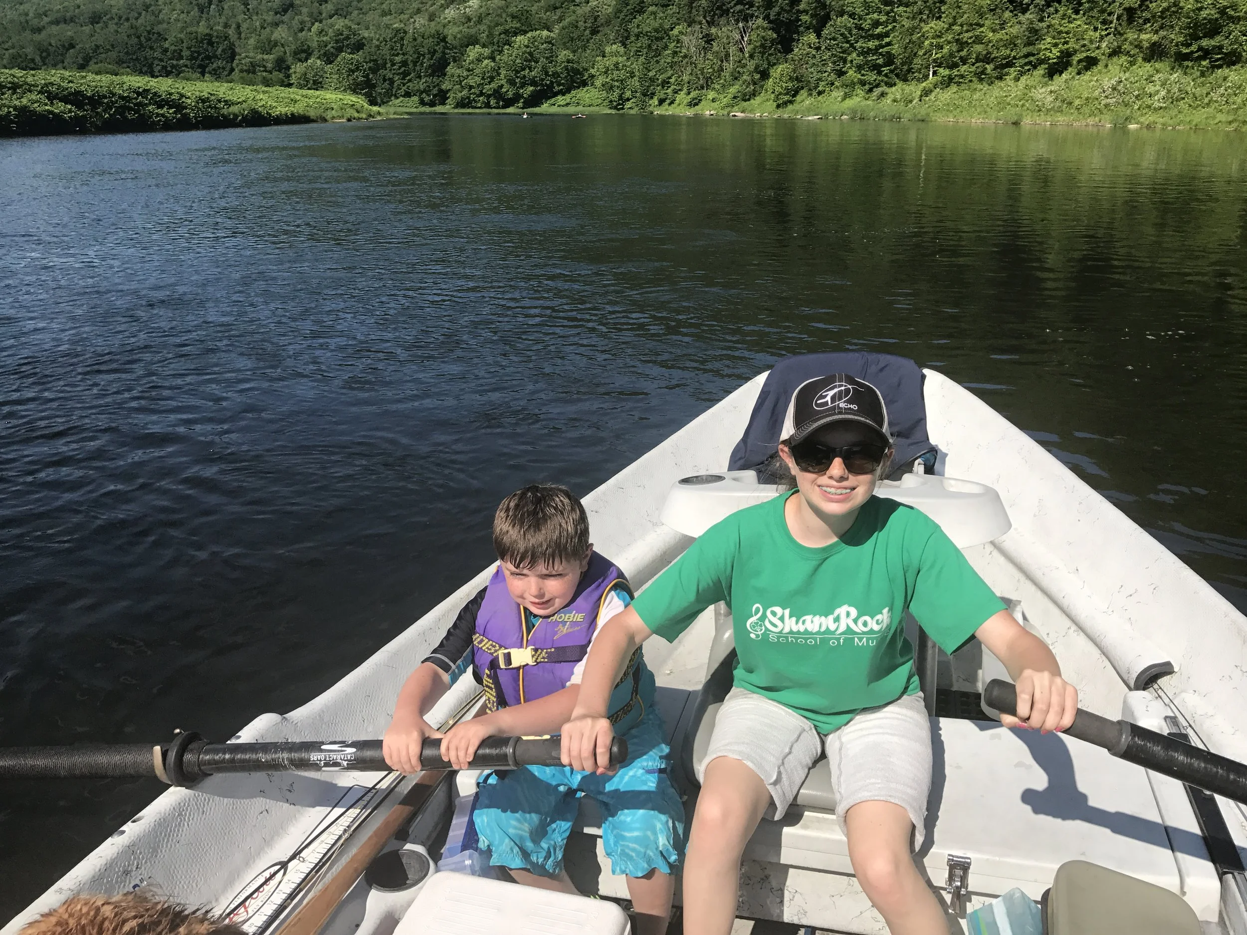 Two children and a young woman on a boat in a river surrounded by green trees. The children are sitting at the front, wearing life jackets and holding an oar. The woman, wearing sunglasses and a green shirt, is smiling and sitting behind them, holding a steering wheel.