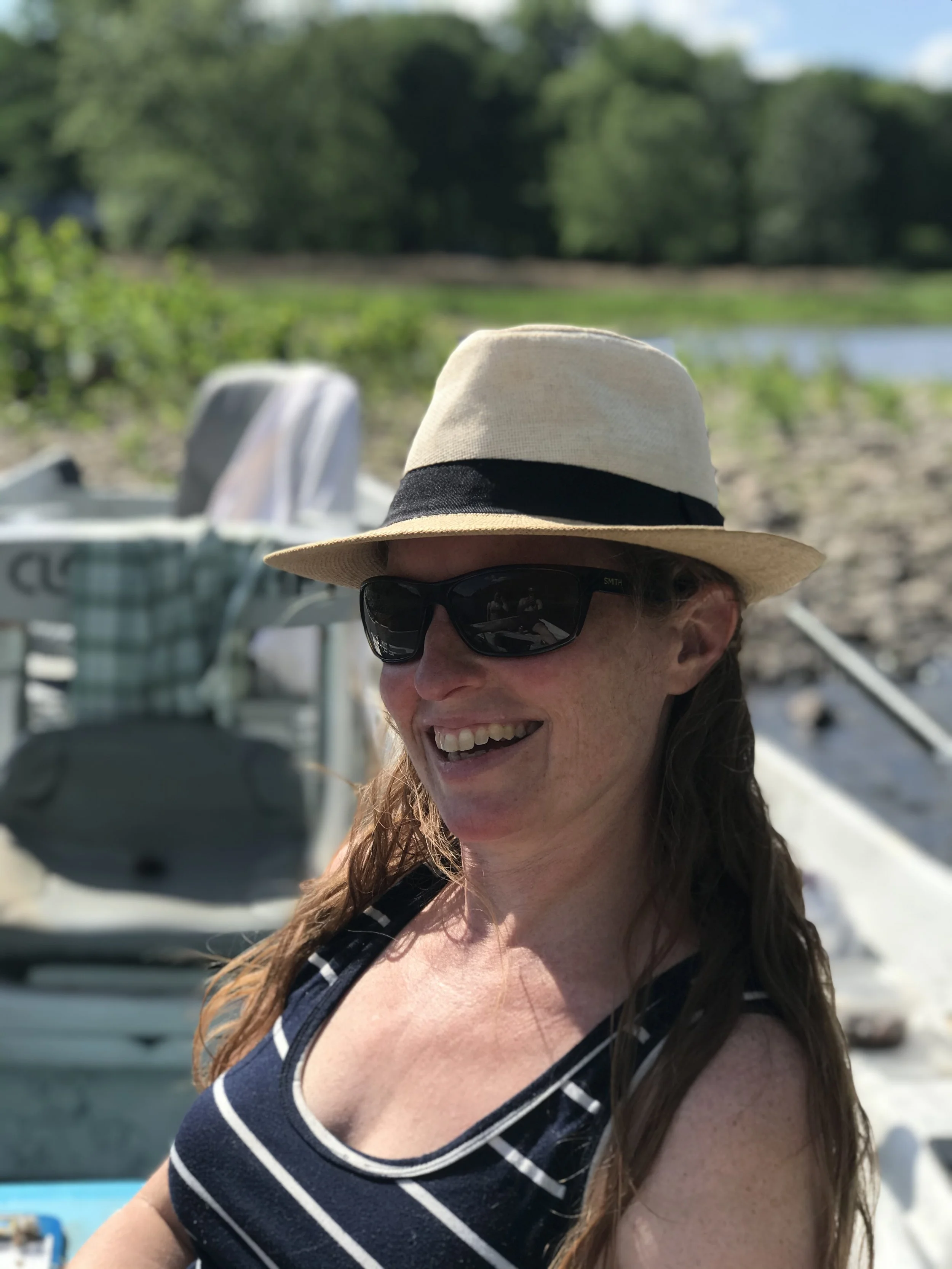 A woman with long brown hair, wearing sunglasses, a wide-brimmed straw hat with a black band, and a navy blue and white striped sleeveless top, smiling outdoors on a sunny day near a body of water with green trees in the background.