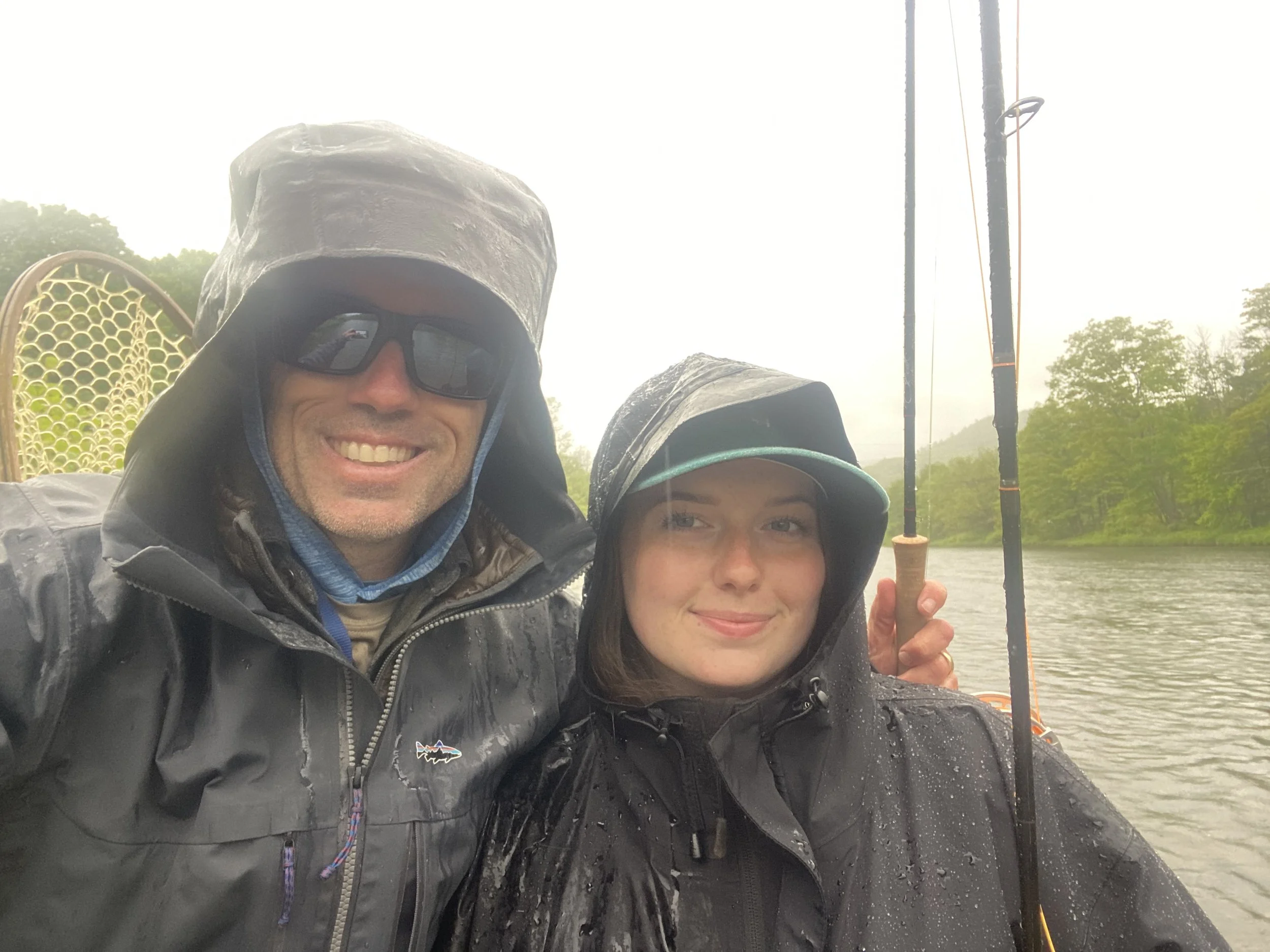 Two people, one male and one female, on a boat during a rainy day, both wearing dark raincoats and hats, with fishing rods visible in the background.