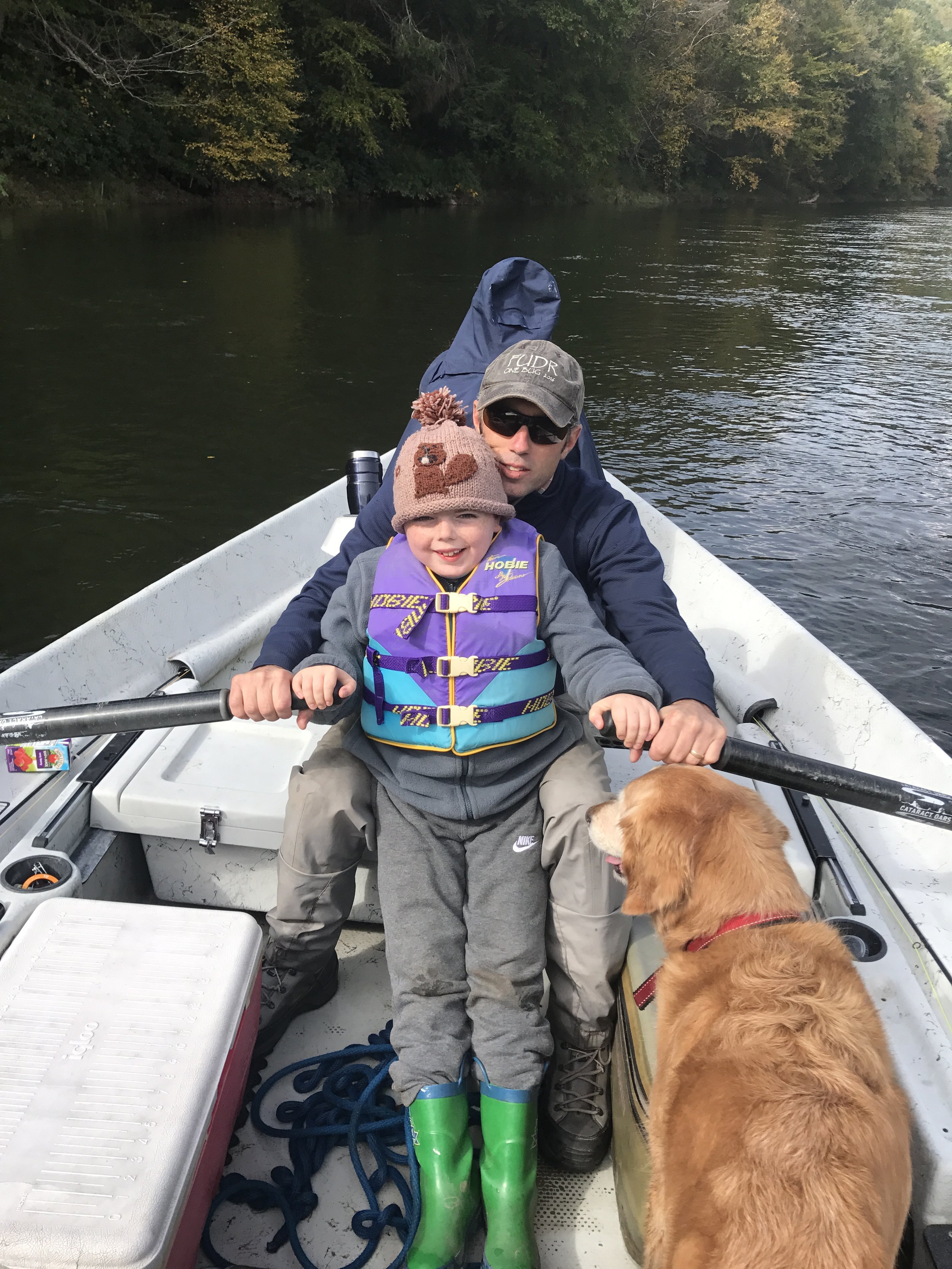 A man and a boy sitting in a small boat on a river, with a dog near the boy. The man is wearing a cap and sunglasses, and the boy is wearing a bear hat with a knit pom-pom. The boy is wearing a life jacket. The boat is surrounded by water and trees with green leaves.