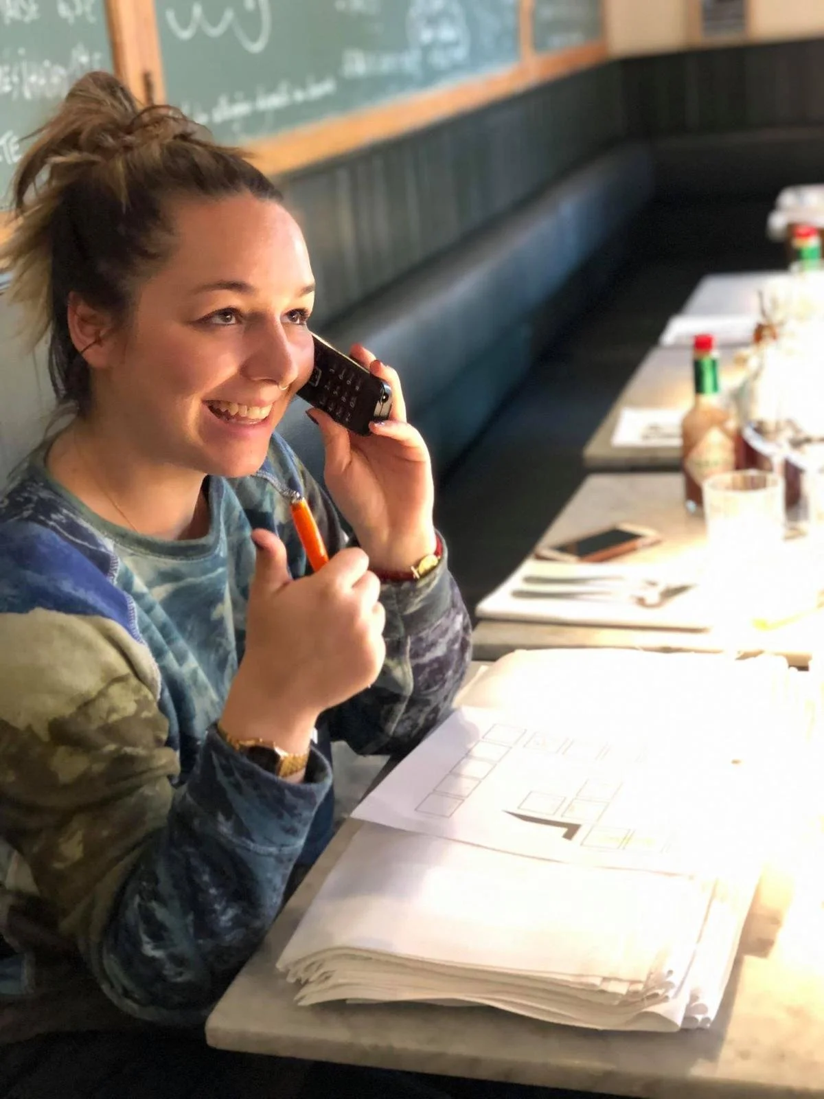 Une jeune femme souriante, portant un sweat camouflage, parle au téléphone dans un restaurant ou un café, avec des papiers et des condiments sur la table.