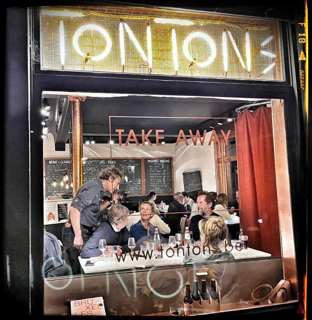 Vue de l'intérieur d'un restaurant ou café vu à travers une vitre, avec des gens assis à une table, un menu visible au fond, et un panneau lumineux avec le mot 'ION' en néon, indiquant la possibilité de plats à emporter. Le site web www.fontons.be est aussi écrit sur la vitre.