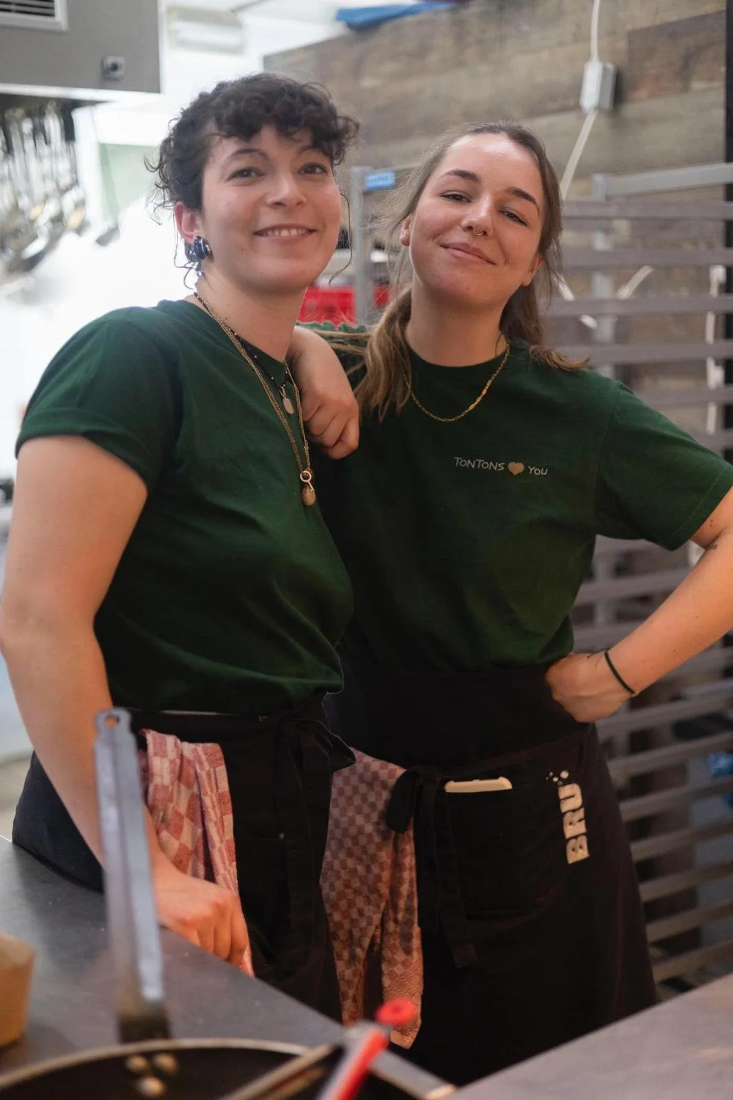 Deux jeunes femmes souriantes portent des t-shirts verts, l'une avec un tablier à carreaux, dans une cuisine
