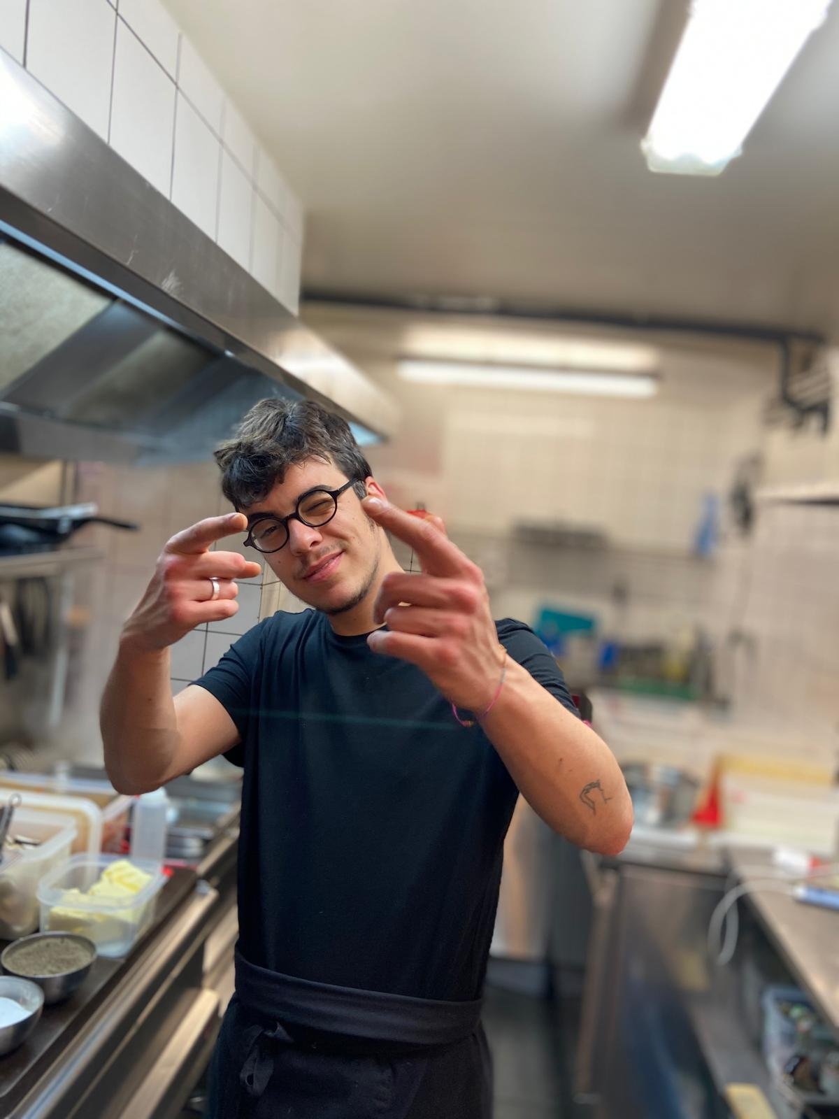 Un jeune homme avec des lunettes, souriant, pointant ses doigts vers la caméra dans une cuisine professionnelle.