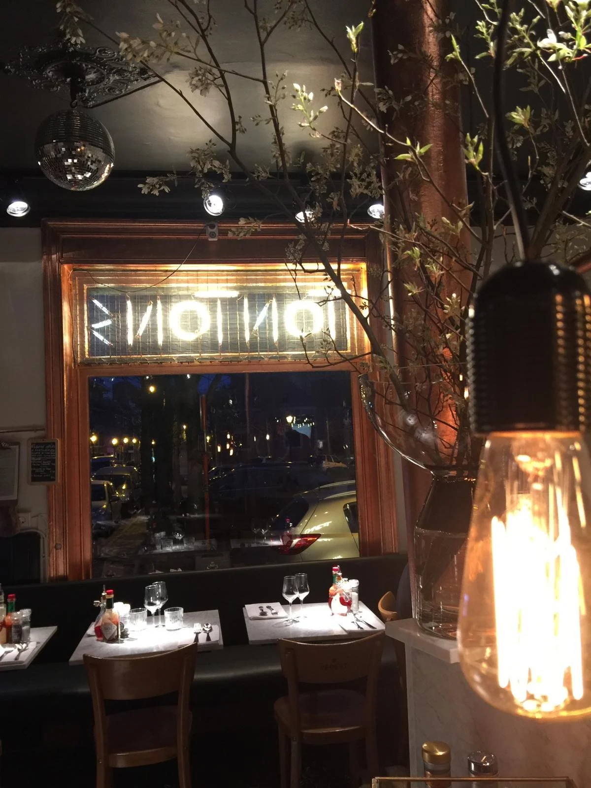 Intérieur d'un restaurant avec une table dressée, un lampadaire avec une ampoule visible, et une grande vase avec des branches. La vue à travers la fenêtre montre une rue avec des voitures la nuit, avec un néon lumineux affichant un mot endommagé ou inversé.